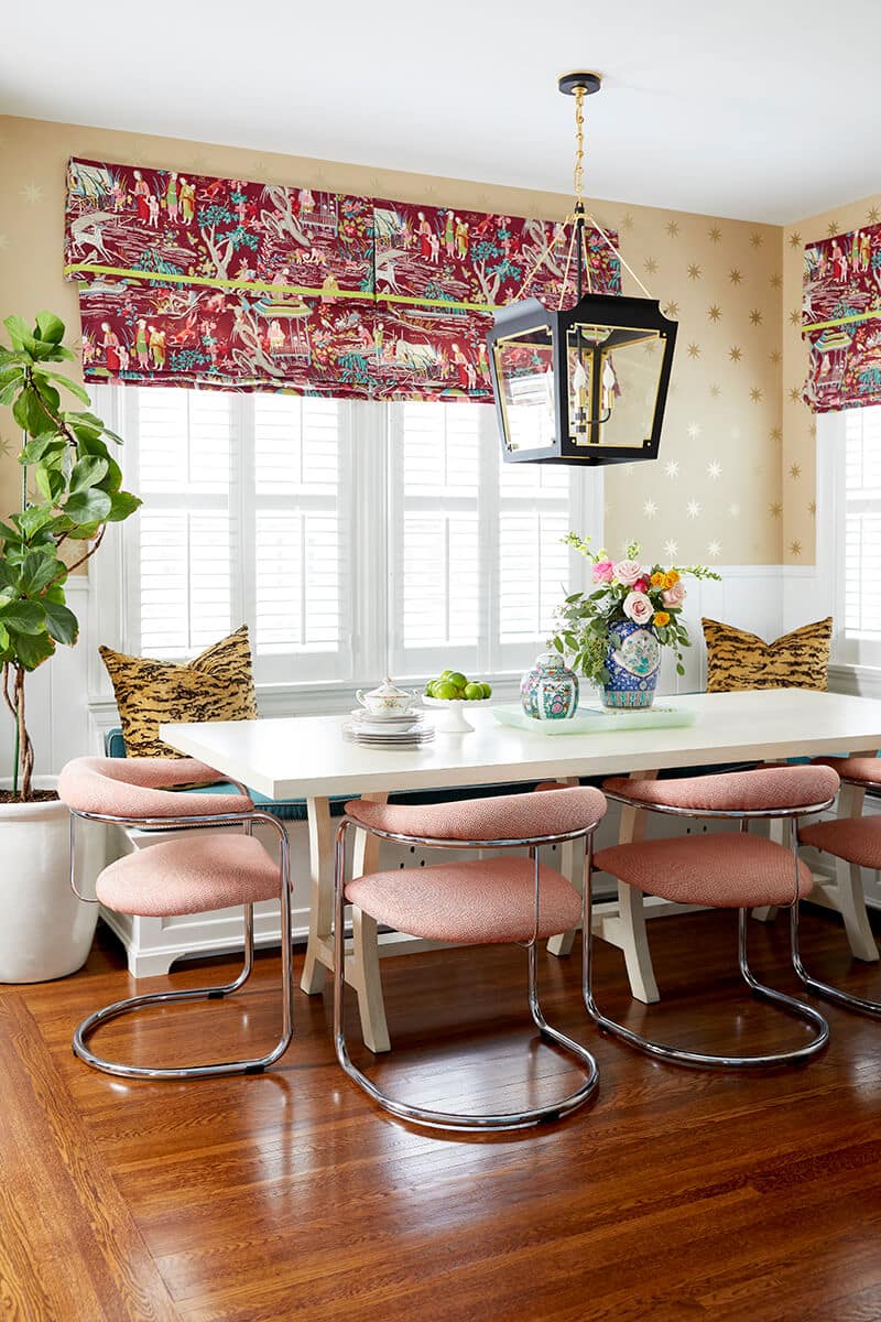 A cozy kitchen breakfast nook is pictured featuring modern pink chairs with a polished silver finish. A bench seat is pictured on the opposite end of the table, with custom-made upholstery in a deep velvet fabric that is comfortable to sit on and beautiful to look at. Cincinnati Interior Designer Laney Reusch's personal style pops out with the tiger-stripe pillows placed on either end of the bench. Above the table is a black pendant light and fun patterned window treatments. This is just a piece of Laney Reusch's own Hyde Park home.